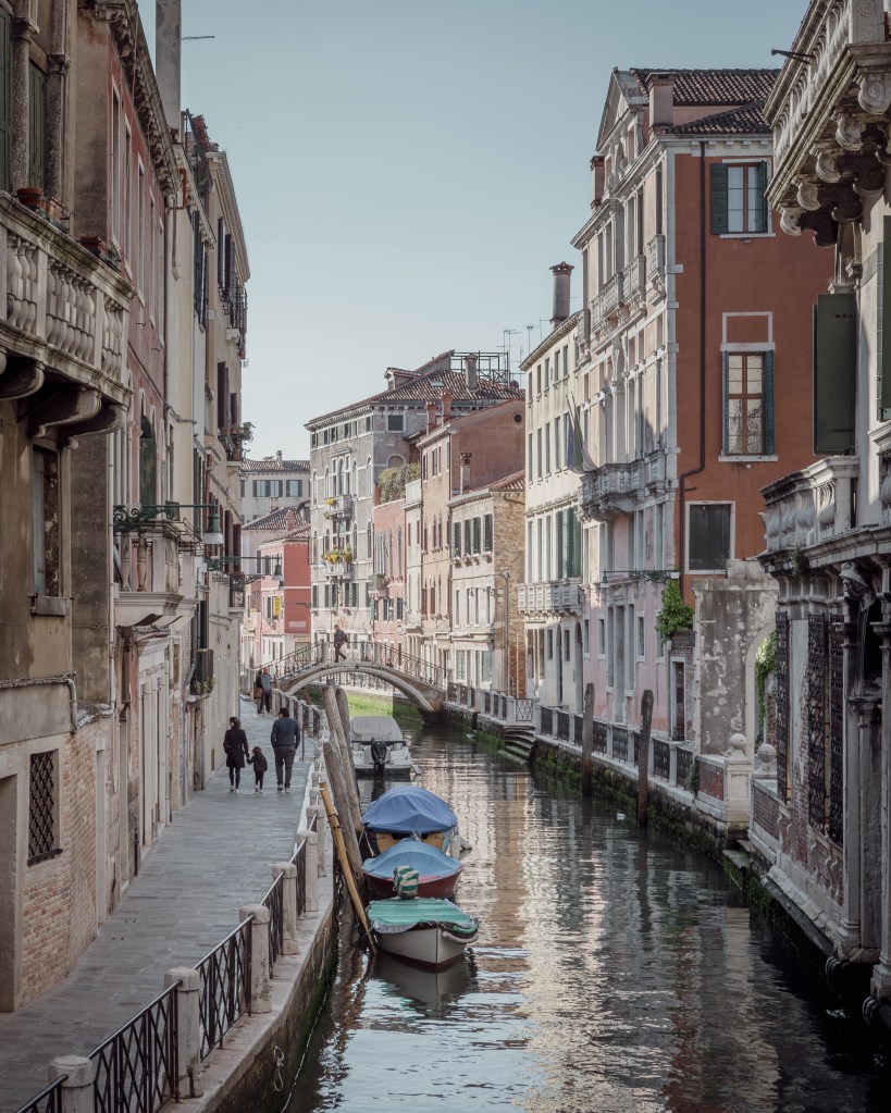 A Venetian canal with parked boats and a family walking along beside the water.