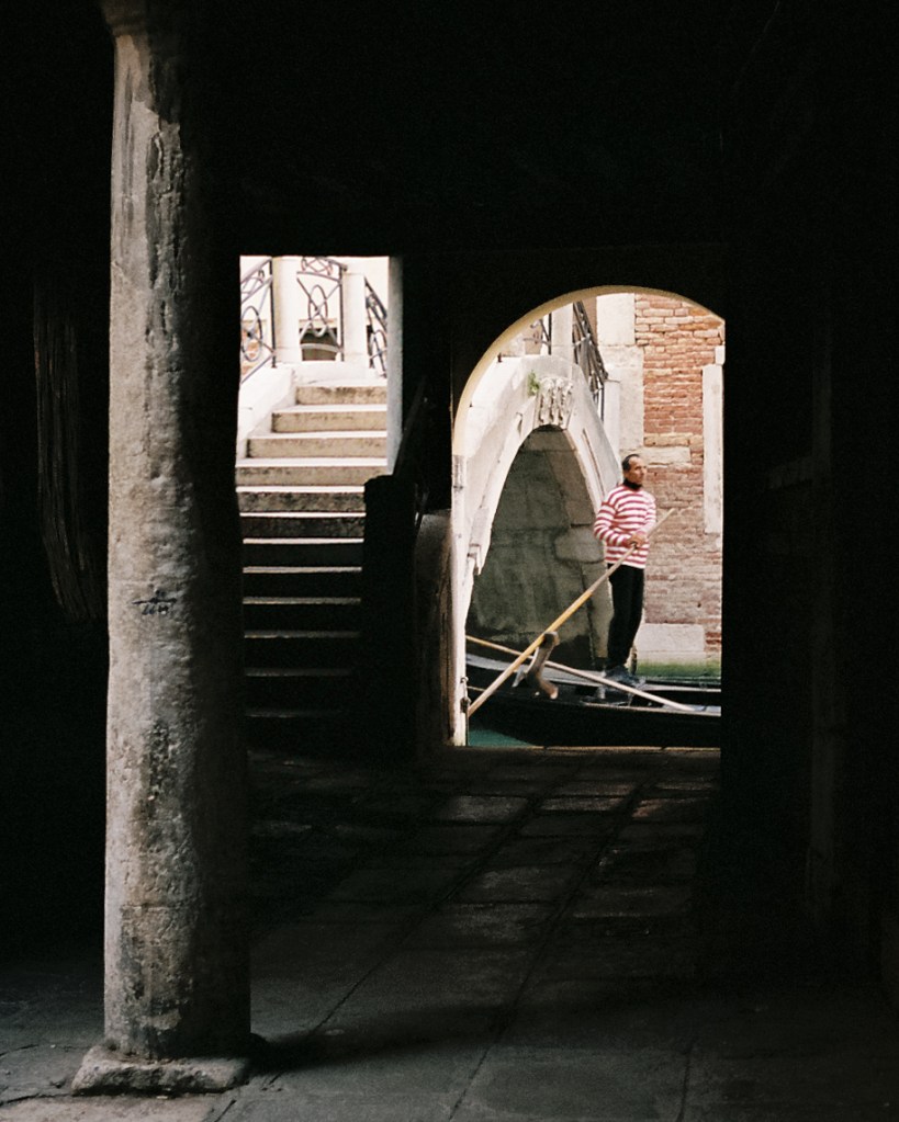 A gondola driver piloting a boat through one of Venice's many canals, framed by an archway.